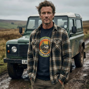 Man in plaid shirt and graphic tee standing by green off-road vehicle in muddy field
