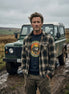 Man in plaid shirt and graphic tee standing by green off-road vehicle in muddy field