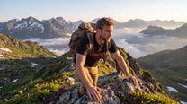 A man hiking on a mountain ridge with a scenic alpine landscape, including snow-capped peaks and clouds, wearing a black t-shirt with orange text and a brown backpack.