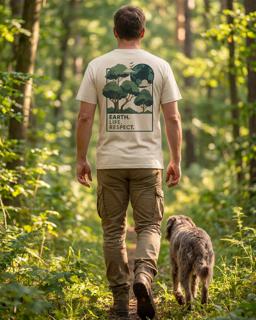 A man seen from behind walking on a forest path with a dog, wearing a beige graphic t-shirt with a tree design and the text 'EARTH LIFE RESPECT'.