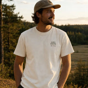 A man wearing a cream-colored crewneck t-shirt with a small black graphic on the left chest, paired with a brown baseball cap and dark pants, standing outdoors in a field during golden hour.
