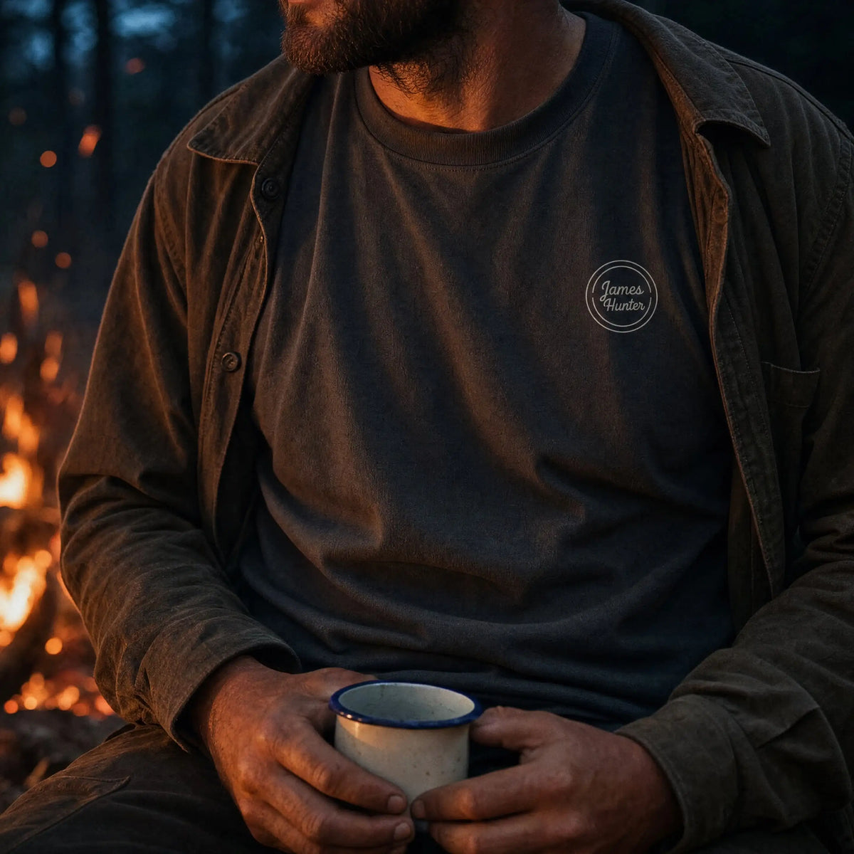 A man wearing a dark charcoal gray t-shirt with a small white circular 'Horizon Wonders' logo on the left chest, layered under a brown overshirt, holding a white enamel mug with a blue rim in front of a warm campfire.