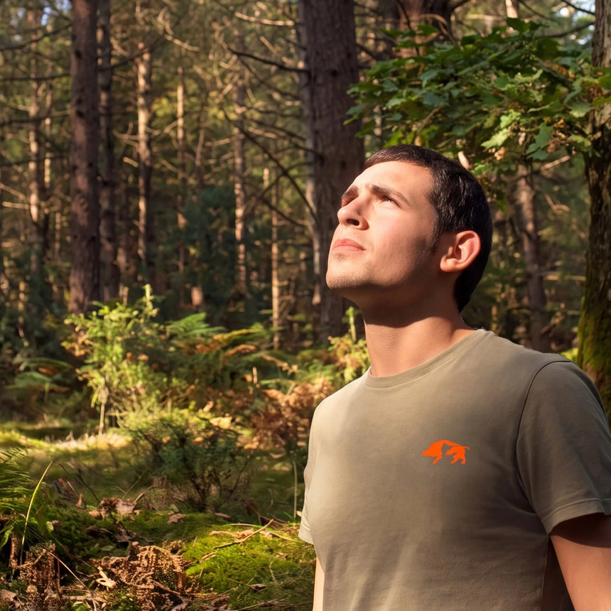 A man wearing a muted olive green t-shirt with a small orange logo on the chest stands in a sunlit pine forest.