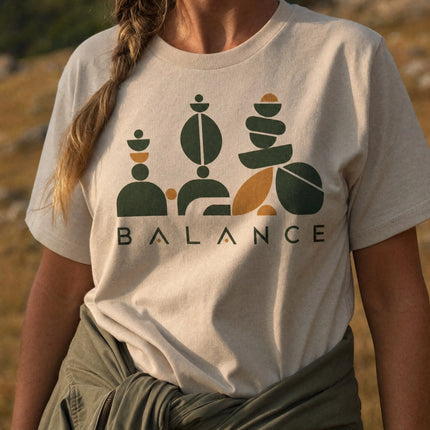 A person outdoors wearing a light beige graphic T-shirt with stacked geometric shapes in dark green and mustard yellow above the word 