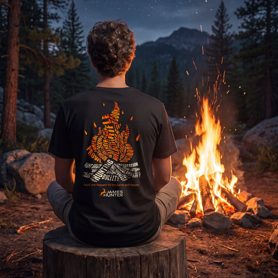 A person sits on a tree stump facing a glowing campfire at night in a forest, wearing a black T-shirt with a large orange and white graphic print on the back. Mountains and a starry sky are visible in the background.