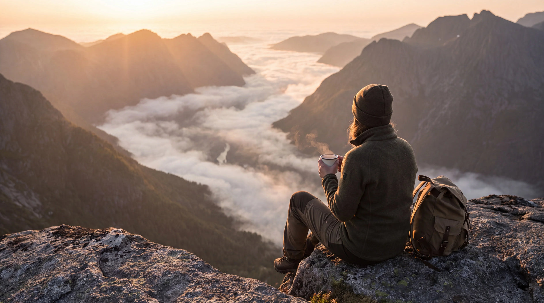A person sitting on a rocky mountain peak overlooking a misty valley at sunrise, with mountains, clouds, and a backpack beside them.