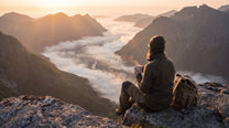 A person sitting on a rocky mountain peak overlooking a misty valley at sunrise, with mountains, clouds, and a backpack beside them.