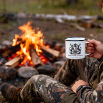 Person in camouflage clothing holding a mug with a logo by a campfire in nature.