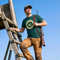 Man on a wooden ladder wearing a green t-shirt with a logo and text, holding a rifle against a clear blue sky.