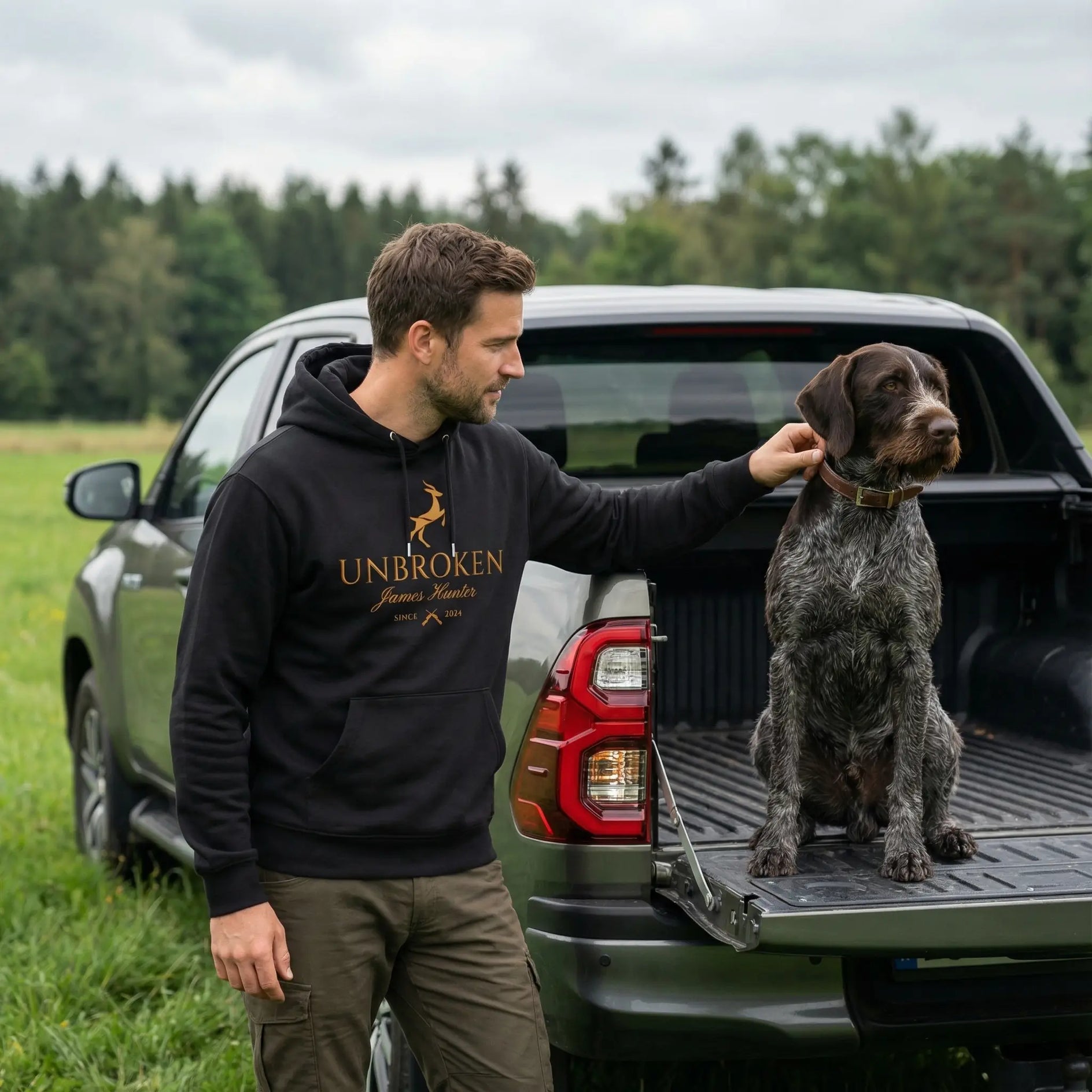 Man in a 'Unbroken' hoodie with a dog on a truck bed in a field