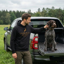 Man in a 'Unbroken' hoodie with a dog on a truck bed in a field
