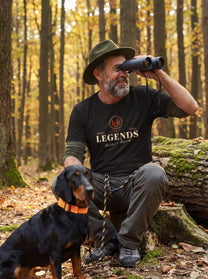 Man in a forest wearing a 'Legends' t-shirt with binoculars and a dog.