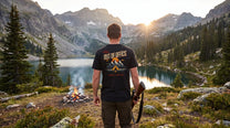Man standing by a campfire with mountains and lake in the background