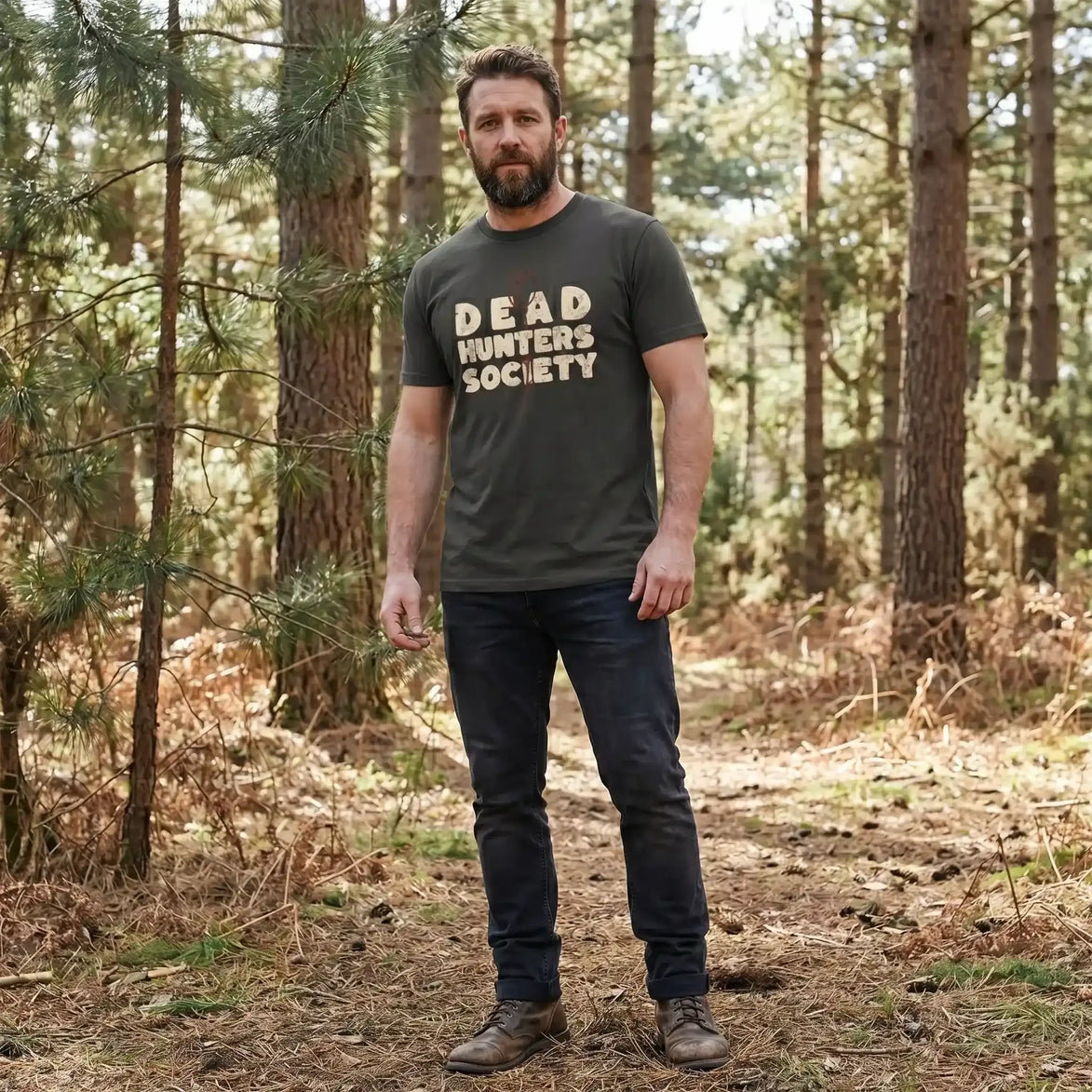 Man in forest wearing olive 'Dead Hunters Society' tee, eco-friendly outdoor apparel for nature enthusiasts.