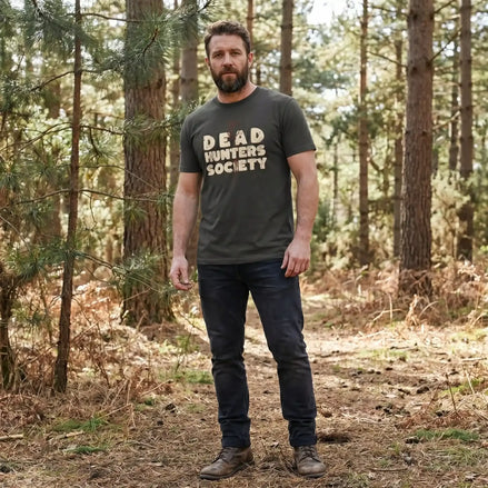 Man in forest wearing olive 'Dead Hunters Society' tee, eco-friendly outdoor apparel for nature enthusiasts.