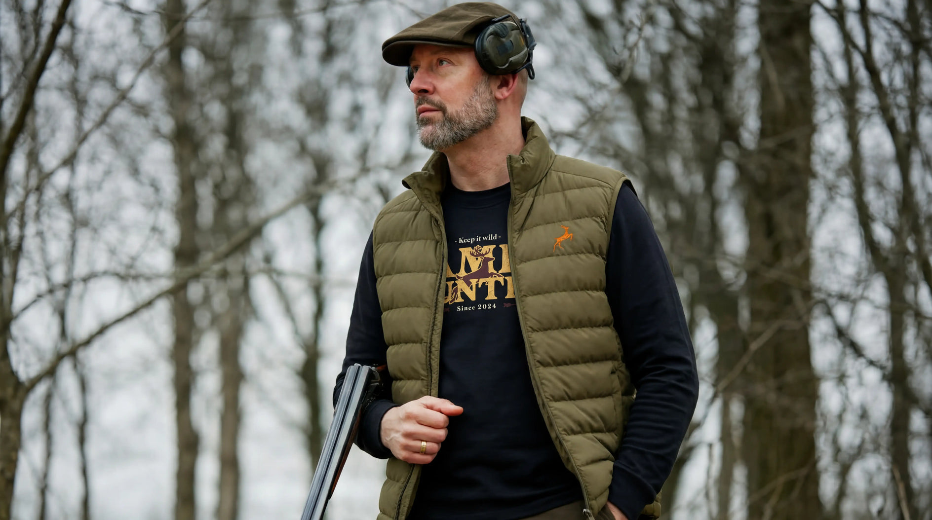 Man wearing a green vest over a black t-shirt with text, standing in a forest.