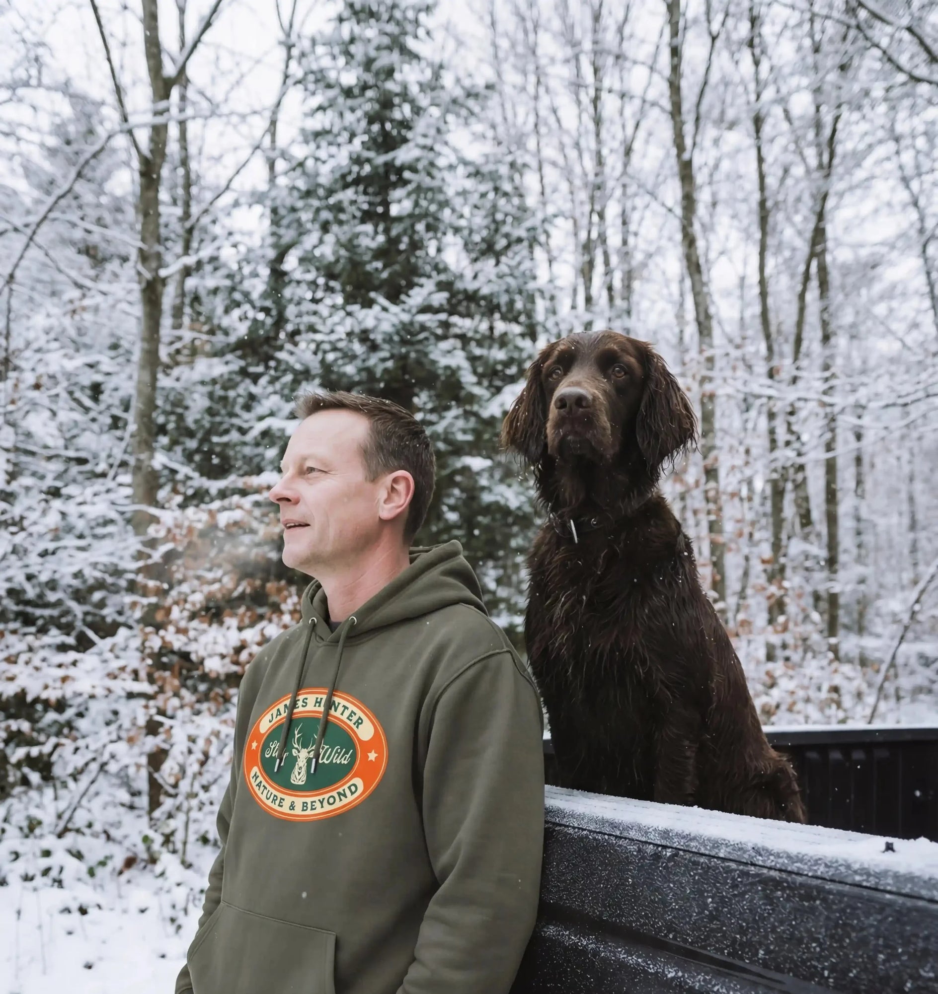 Man wearing a green hoodie with a logo standing next to a truck with a dog in the snow.