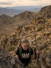 Woman hunter in rugged mountains wearing 'Dead Hunters Society' tee, holding rifle, eco-friendly outdoor apparel