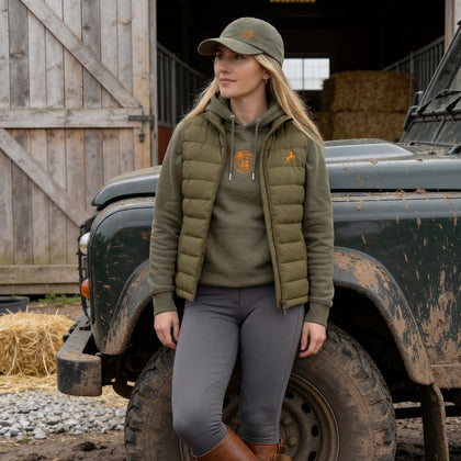 Woman in green jacket and cap standing next to a rustic vehicle in a barn setting